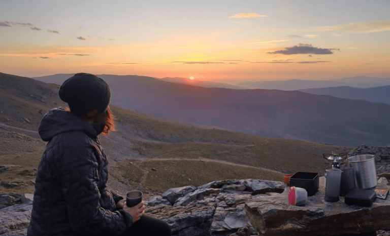 ana explora disfrutando un café caliente con una AeroPress Go al amanecer en la montaña, a casi 3000 metros de altura, con equipo de camping sobre una roca y vistas panorámicas de Sierra Nevada